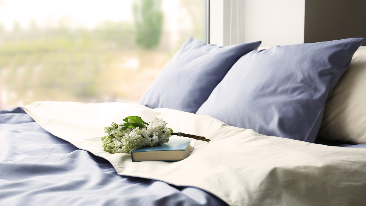 A cozy bed with light-blue pillows and matching bedding sits by a large window. A book and white flowers rest on the duvet, adding a serene touch.