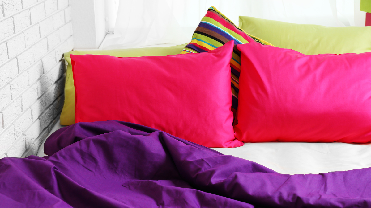 A vibrant bed setup featuring bold pink pillows, a purple blanket, and a mix of striped and yellow cushions creates a lively and colorful atmosphere. Perfect for adding energy to any room.