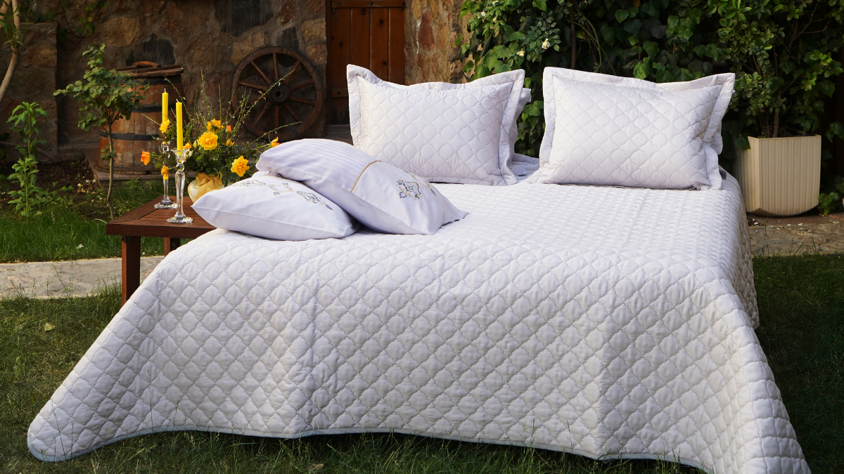 An outdoor setup featuring a neatly made bed with quilted white bedding and pillows, surrounded by greenery. A small wooden table with yellow flowers and candles adds a cozy, rustic charm to the scene.