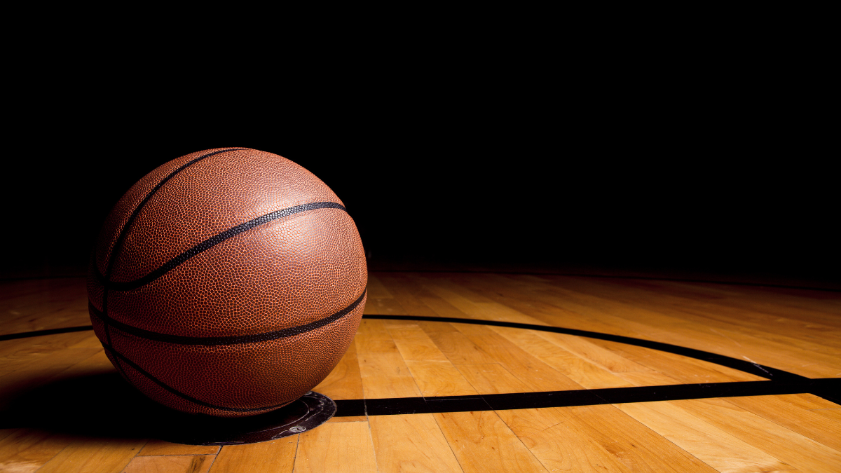 A classic basketball resting on a wooden court, illuminated dramatically against a dark background. The court lines subtly frame the ball, creating a sense of focus and anticipation.
