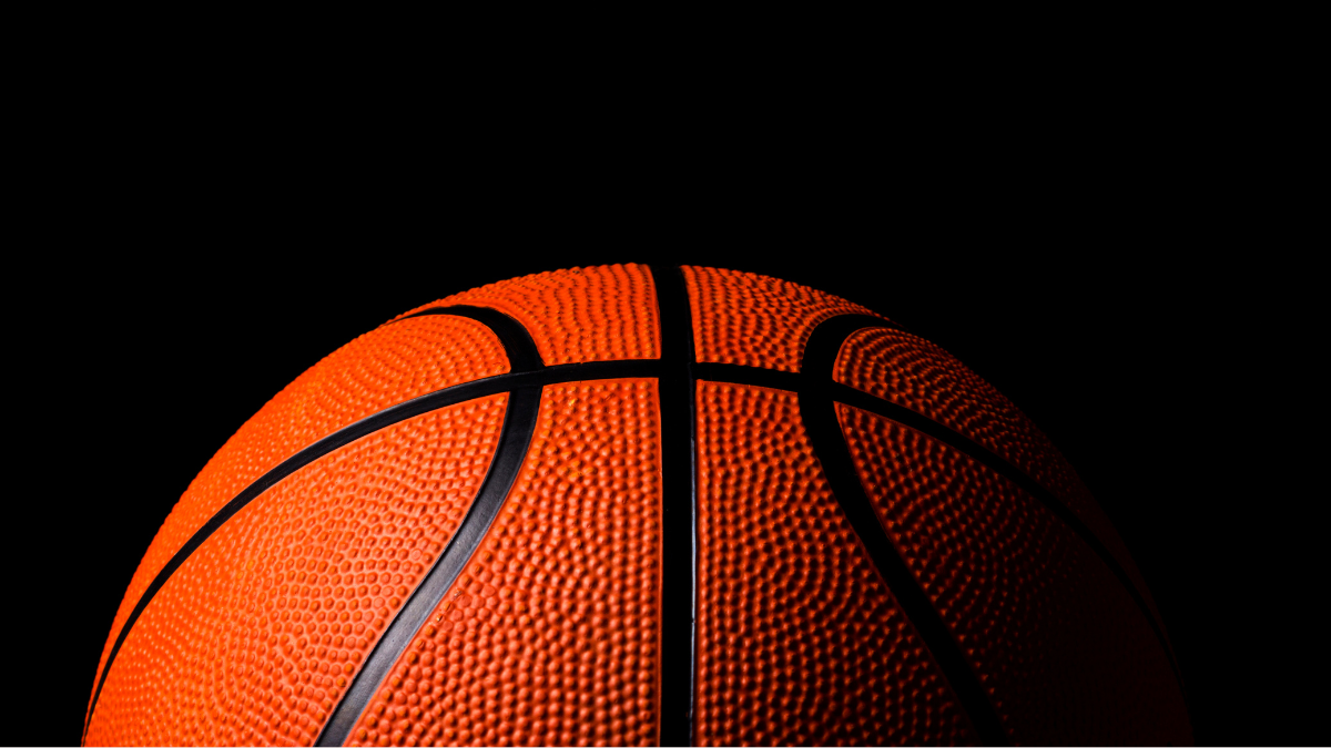 A close-up of a basketball against a deep black background, highlighting the textured surface and distinctive black lines. The contrast emphasizes the ball's vivid orange color and detailed grain pattern.