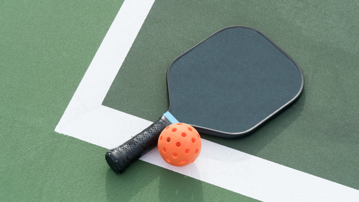 This image showcases a pickleball paddle and an orange perforated ball resting on the corner of a green outdoor court, highlighting the simplicity and accessibility of the sport.