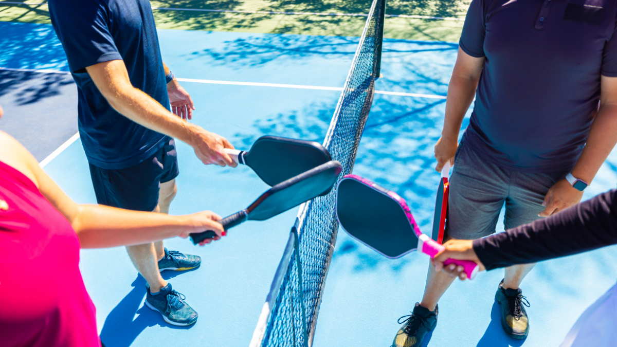 This image captures a sportsmanlike moment as four players tap their pickleball paddles over the net on a vibrant blue outdoor court, symbolizing teamwork and friendly competition.