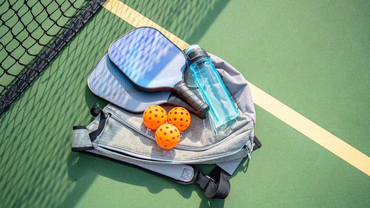This image features essential pickleball gear, including a paddle, three orange balls, and a water bottle, neatly placed on a backpack beside a court net. It highlights preparation for an engaging game.