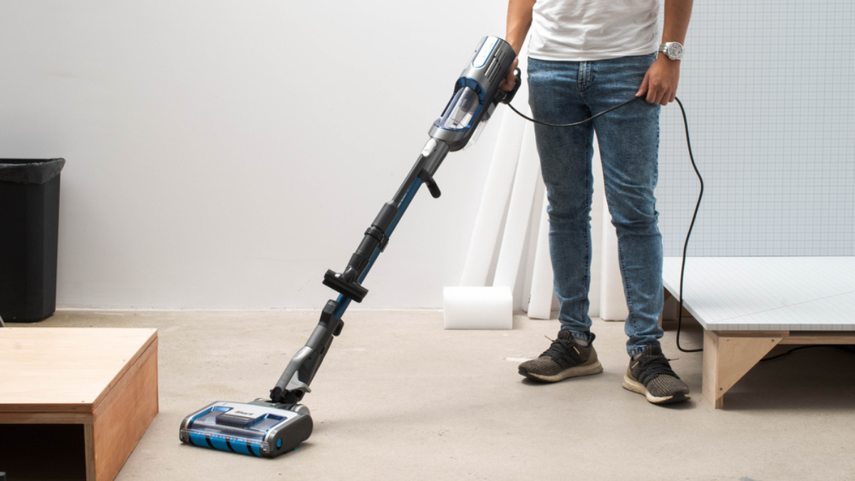 A person using a corded stick vacuum cleaner on a smooth floor surface in a minimal indoor setting.