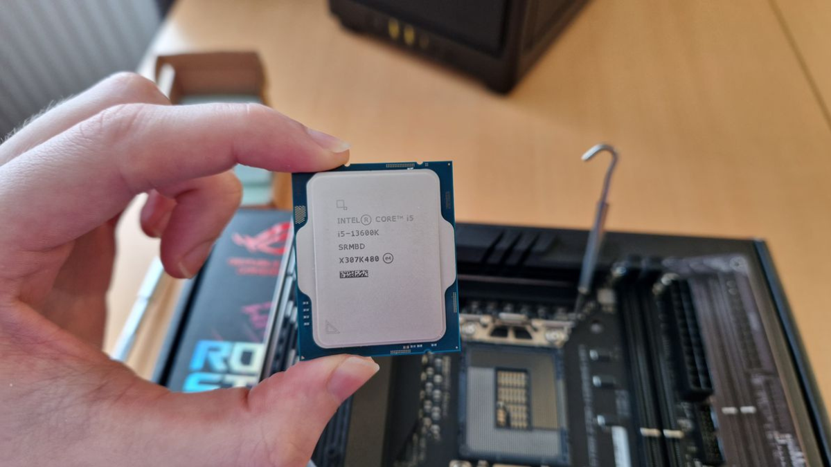 A person holding an Intel Core i5 processor above a motherboard setup on a wooden table.