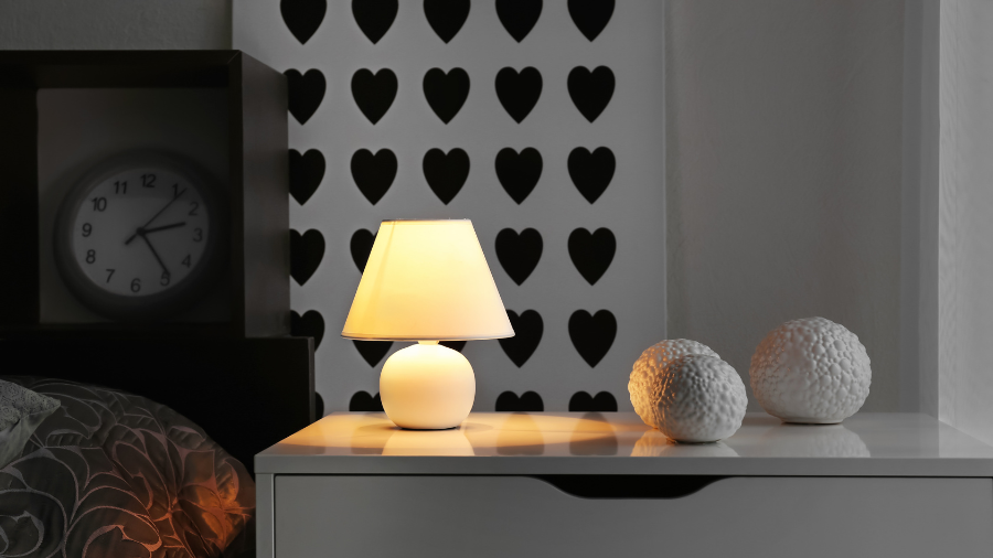 A cozy bedside table setup featuring a glowing round-based table lamp, decorative textured white spheres, and a black-and-white heart-patterned wall in the background.