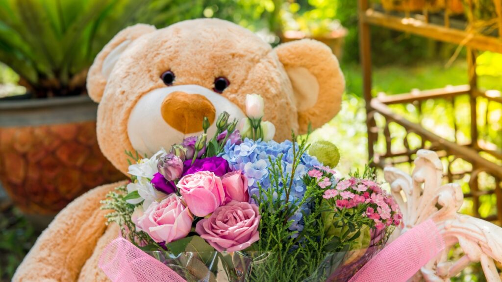 A large teddy bear holding a colorful bouquet of roses and other flowers with a pink ribbon.