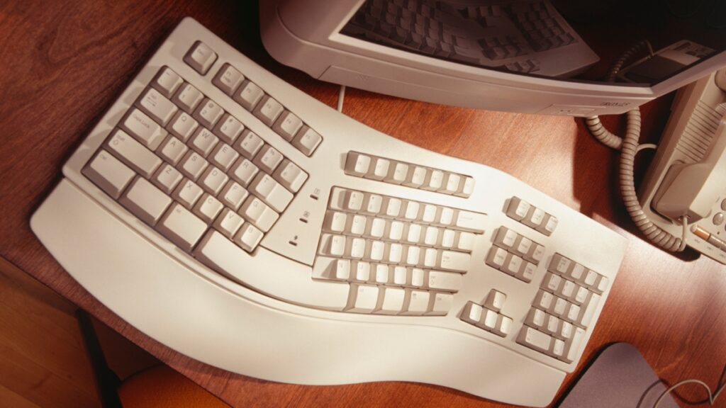 A vintage beige ergonomic keyboard with a split design and built-in wrist rest is placed on a wooden desk near an old computer monitor and telephone.