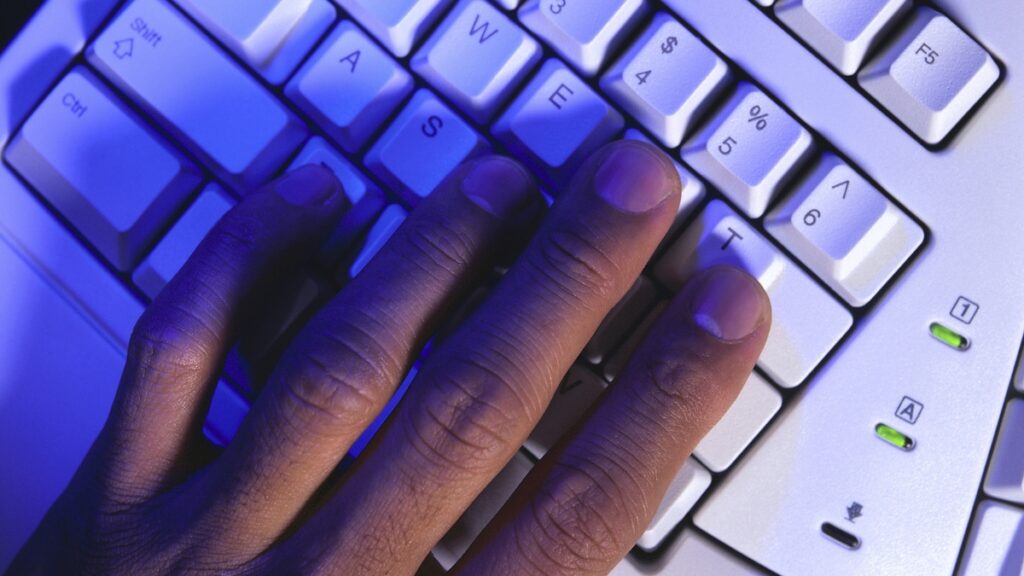A close-up of a hand typing on a white keyboard under blue lighting, with fingers resting on the keys.