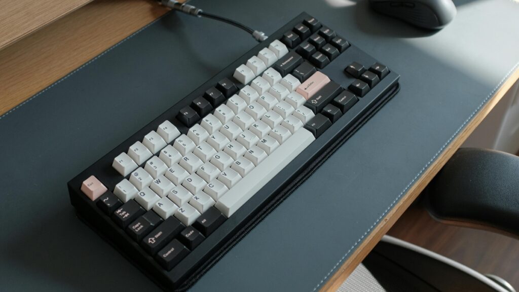 A compact mechanical keyboard with a black and white keycap set, accented with pink keys, sits on a dark desk mat with a connected cable.