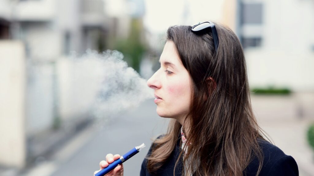 A woman with long brown hair vaping outdoors, exhaling a cloud of vapor while holding a blue vape pen.