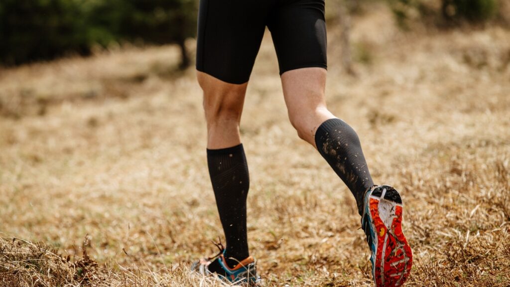 a runner wearing black compression socks and shorts, running on a dry grassy trail with mud on their shoes.
