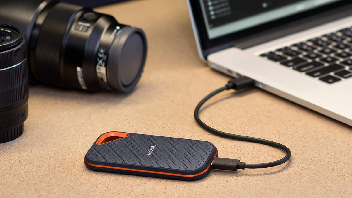 A compact external hard drive connected to a laptop, with a camera lens in the background, suggesting a setup for photographers or content creators needing portable storage.