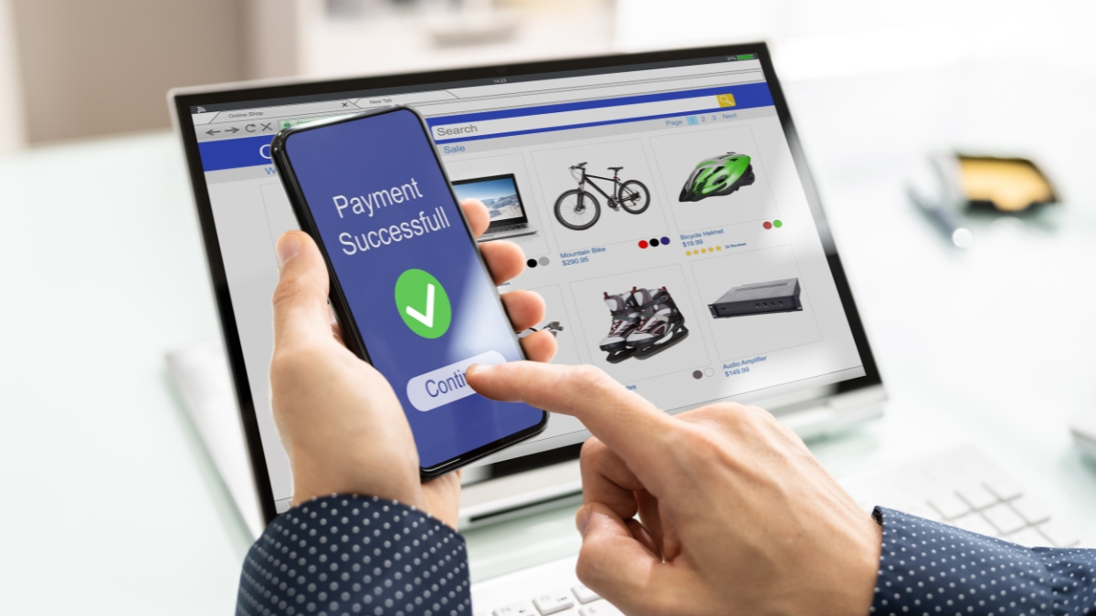 A person completes an online shopping payment on a smartphone while browsing products like bikes and helmets on a laptop screen.