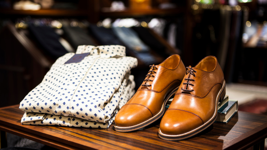 A pair of polished brown leather dress shoes with black laces are displayed on a wooden table next to a neatly folded white dress shirt with a small navy blue pattern.