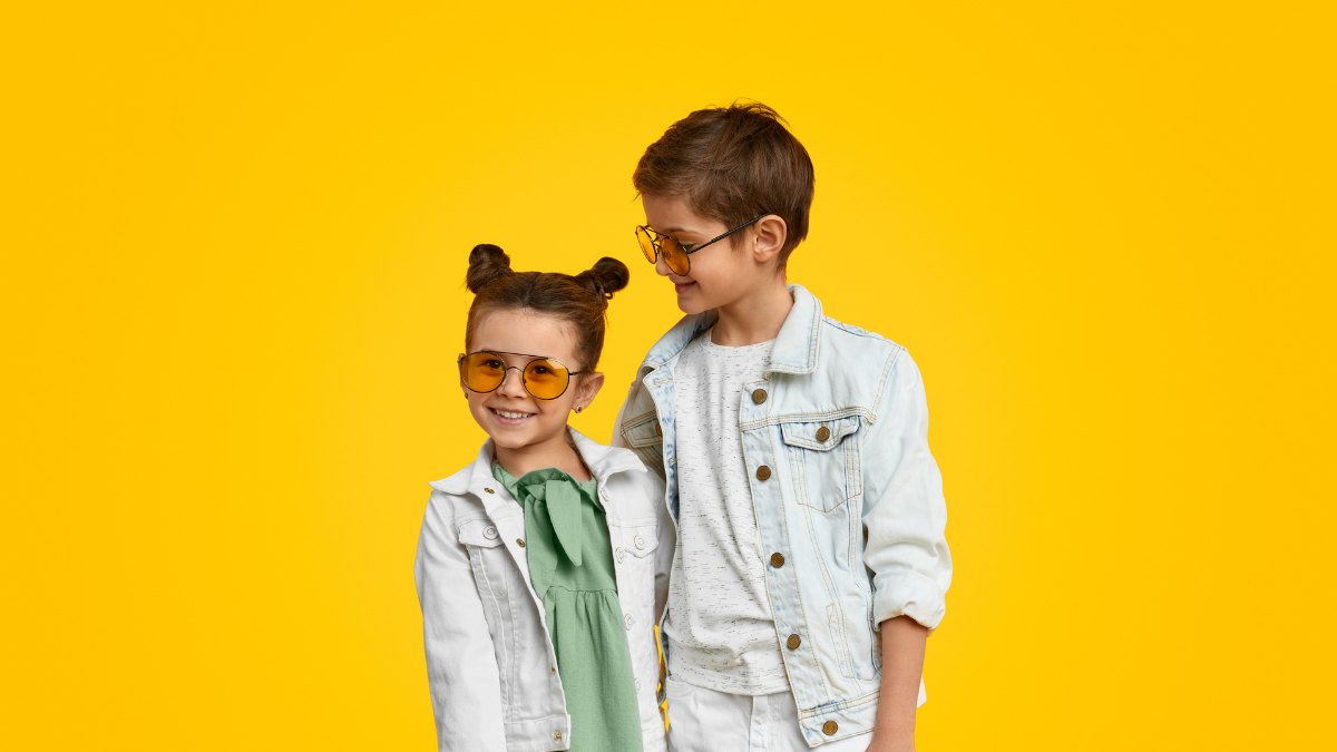 Two stylish young children pose against a vibrant yellow background. They both wear matching light denim jackets and yellow-tinted sunglasses.