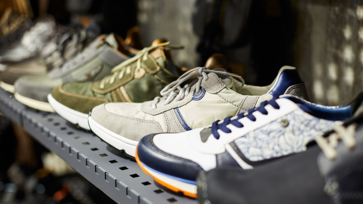 A close-up view of various casual sneakers displayed on a metal shelf. The shoes feature different colors and designs, including green, beige, blue, and white combinations, with distinct stitching and textured materials.