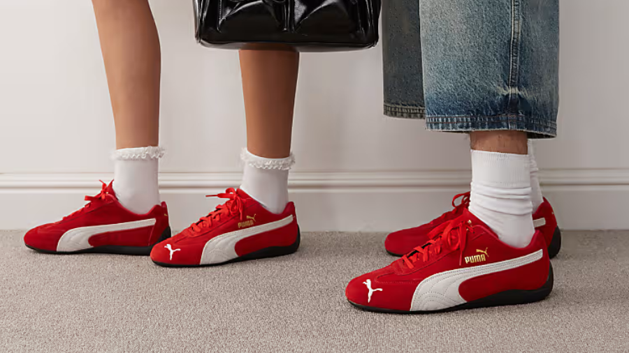 A stylish pair wearing matching red and white Puma sneakers with black soles. One person pairs them with frilly white socks and a black handbag, while the other sports white crew socks and wide-leg denim jeans.