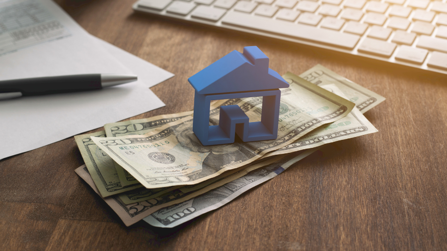 A small blue house-shaped model placed on top of stacked U.S. dollar bills on a wooden desk, with a pen, document, and keyboard in the background