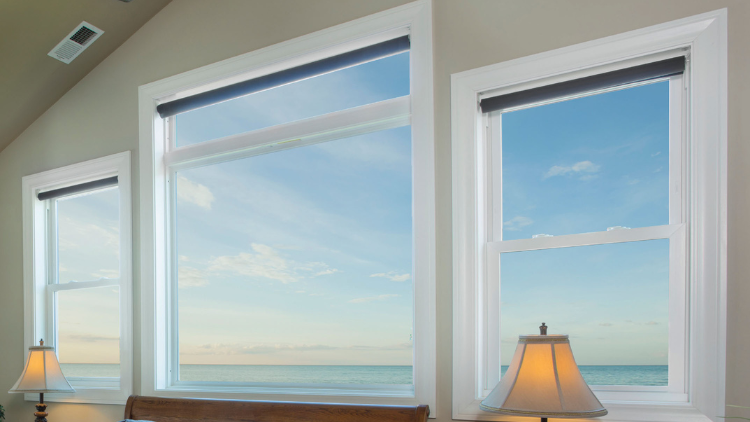 Three large windows with roller shades overlooking a calm ocean view at sunset, framed by soft beige walls and lit by table lamps.