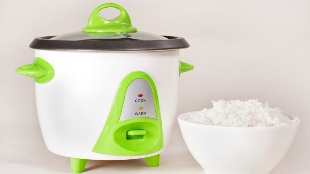 Small electric rice cooker with green handles and a bowl of cooked white rice on a plain background