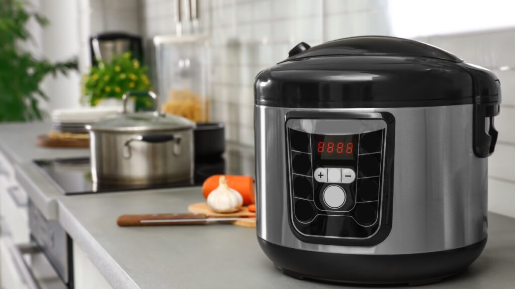 A black stainless steel rice cooker with a digital display on a kitchen counter, surrounded by cooking essentials.