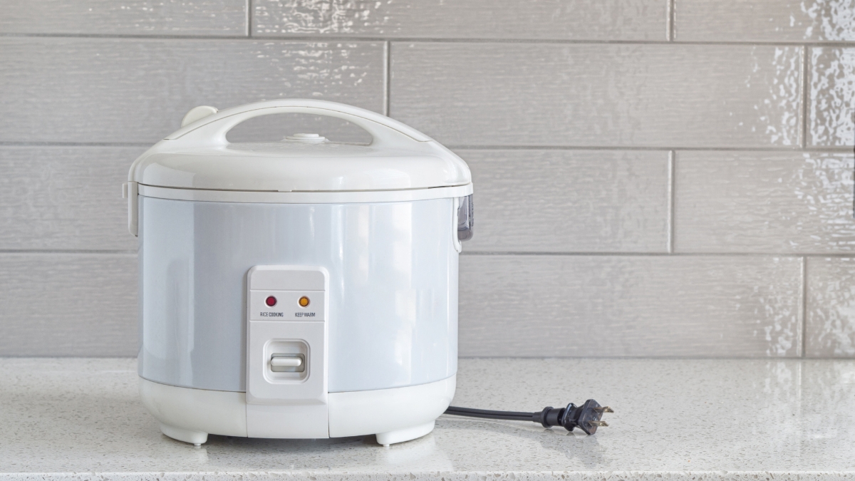 A simple white rice cooker on a speckled countertop with a modern tiled backsplash, plugged out and ready for use.
