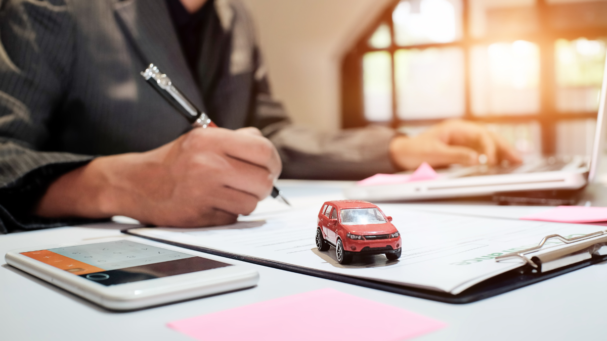 A businessperson signing a car insurance or finance document with a miniature red SUV on top, a calculator, and a laptop on the desk