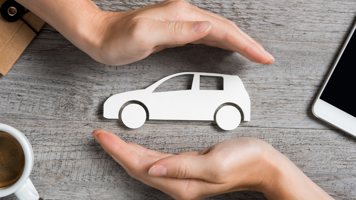 Two hands forming a protective gesture around a white paper cutout of a car on a wooden surface, symbolizing car insurance or safety.