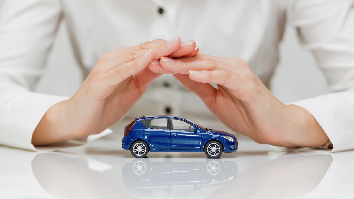 A person in a white shirt cupping their hands over a small blue car model, representing vehicle protection or insurance coverage.