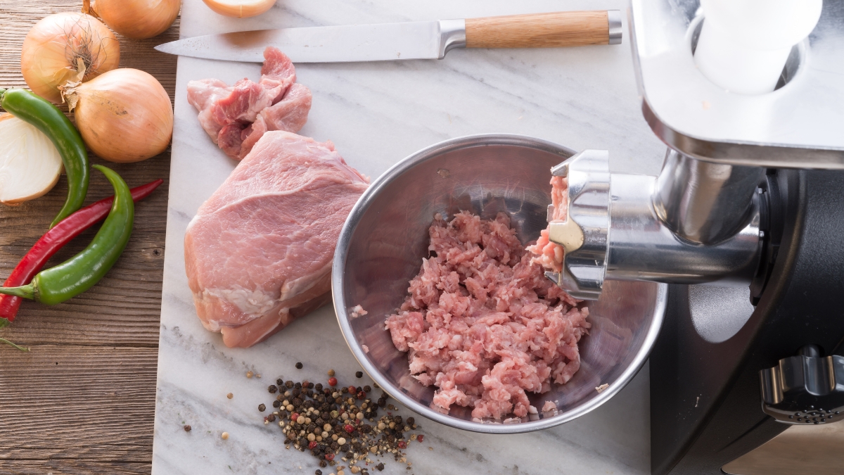 Fresh meat being ground using a stainless steel electric meat grinder with onions, peppers, and spices on a kitchen counter