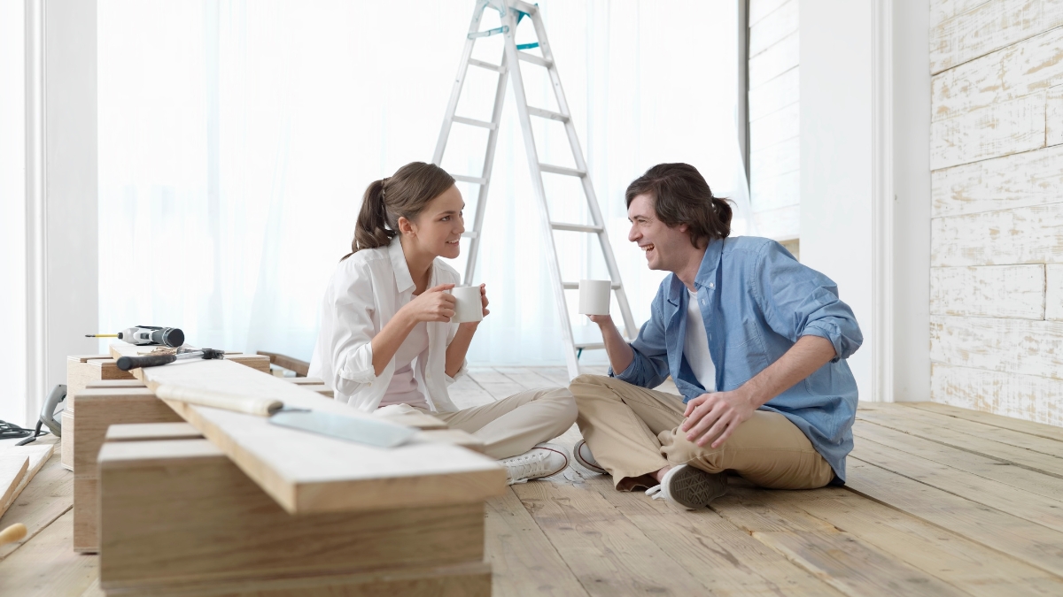 A couple sits on a wooden floor holding coffee mugs while taking a break from home renovation, with tools and a ladder nearby.
