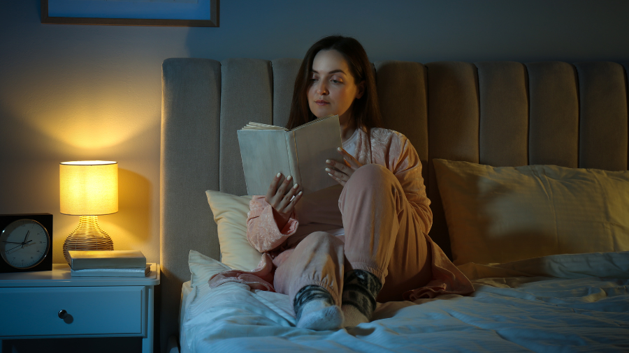 Woman in pink pajamas reading a book in bed at night with a bedside lamp on.