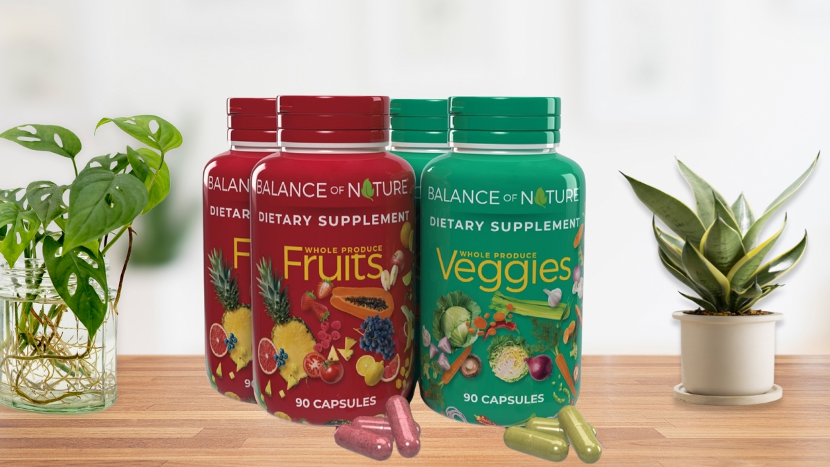 Bottles of Balance of Nature Fruits and Veggies supplements with capsules on a wooden table between two small indoor plants.