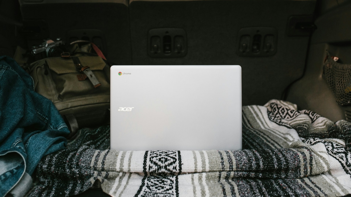 A silver Acer Chromebook sits on a patterned blanket in the back of a car next to a backpack.