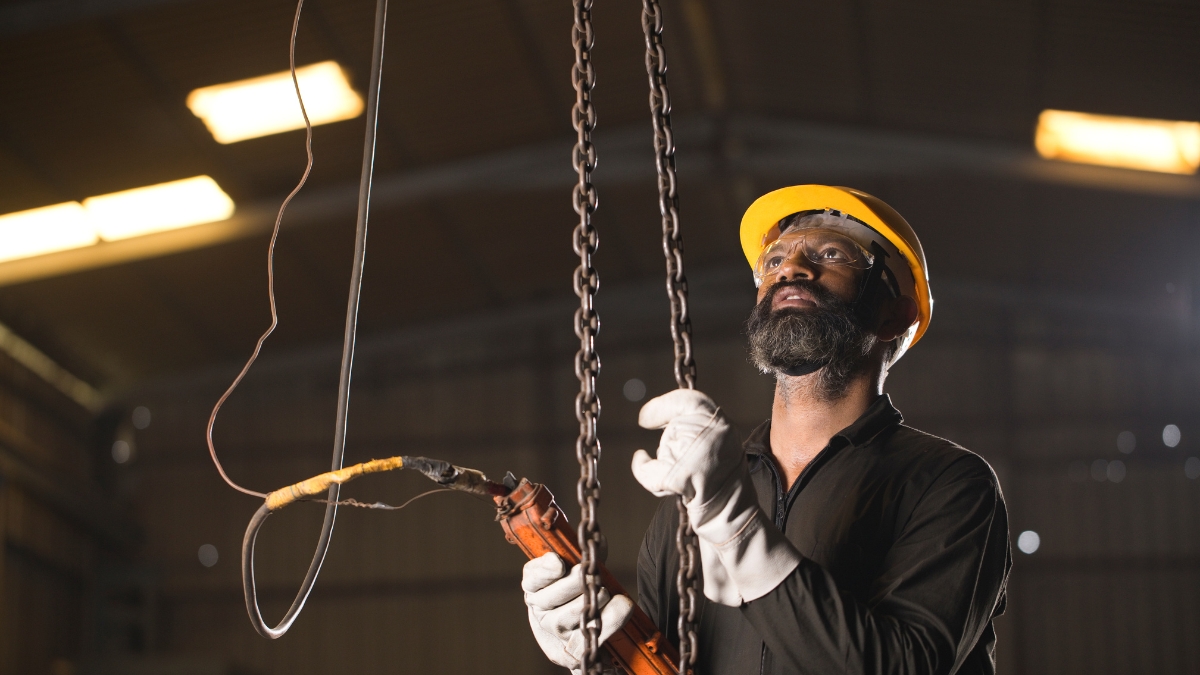 Worker in safety gear operates a chain hoist in a warehouse using a wired control remote.