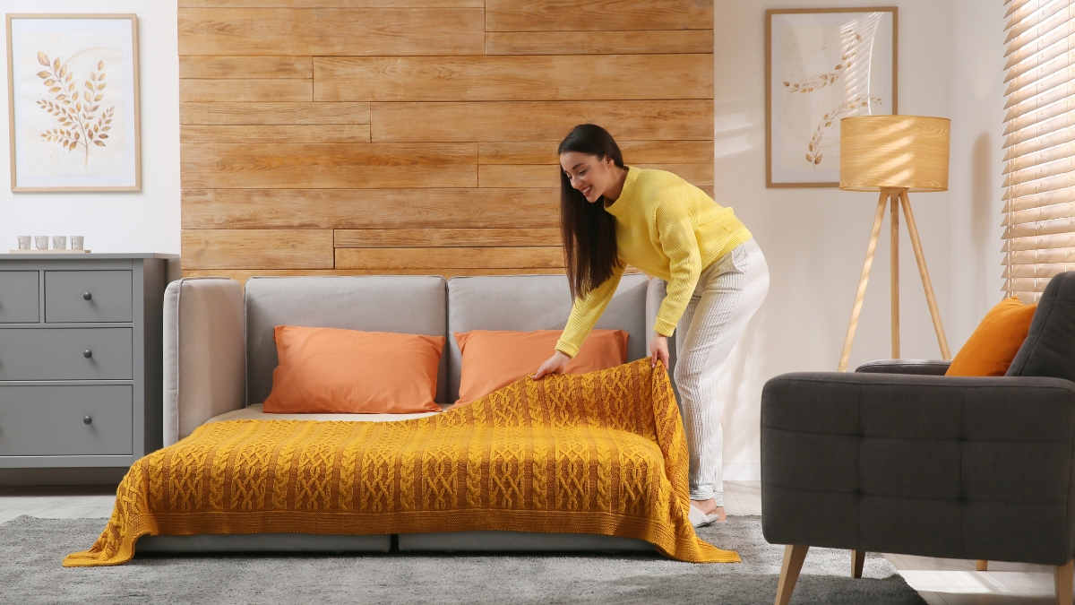Woman spreading a yellow blanket over a pull-out sleeper sofa in a cozy, modern living room.