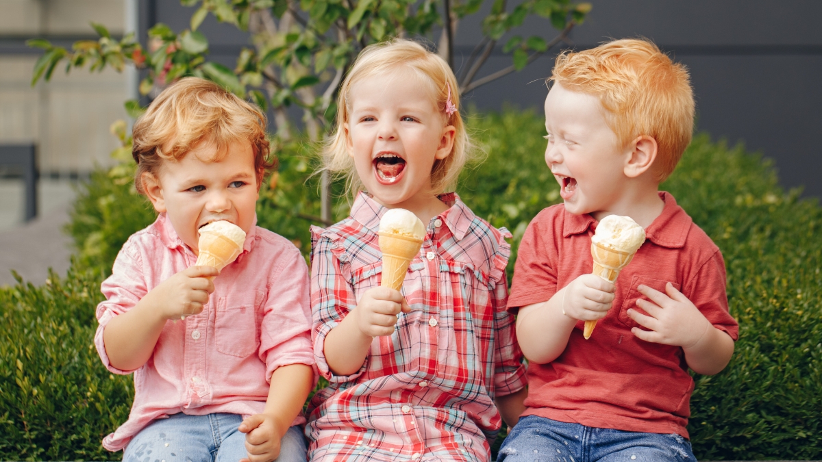 Three happy kids sitting outside, laughing and eating ice cream cones on a sunny day.
