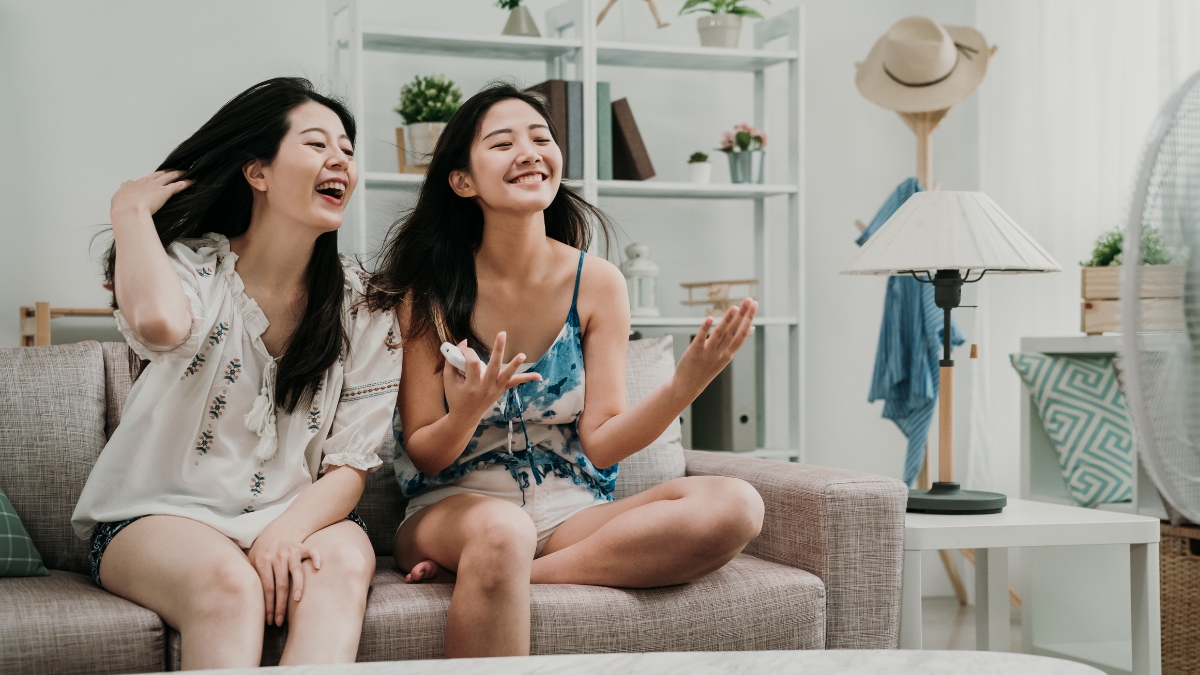 Two women sitting on a couch smiling and enjoying the cool air from a fan or air conditioner on a hot day.