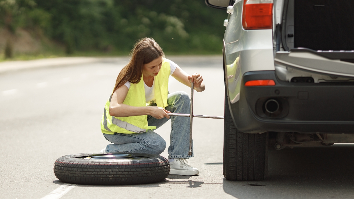 A woman in a safety vest changing a flat tire on the side of the road using a lug wrench.